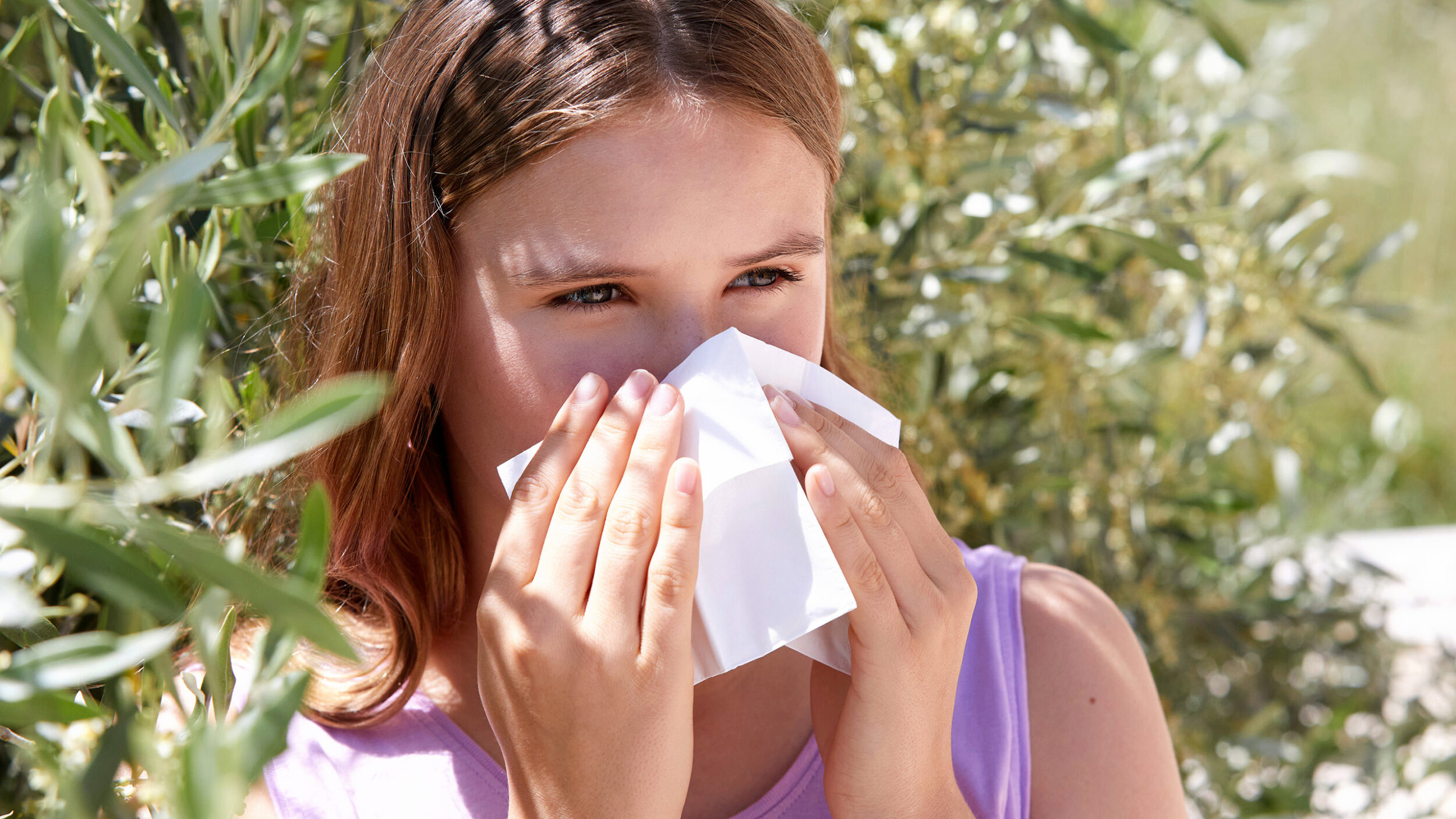 Young woman with seasonal allergy symptoms holding a tissue outdoors.
