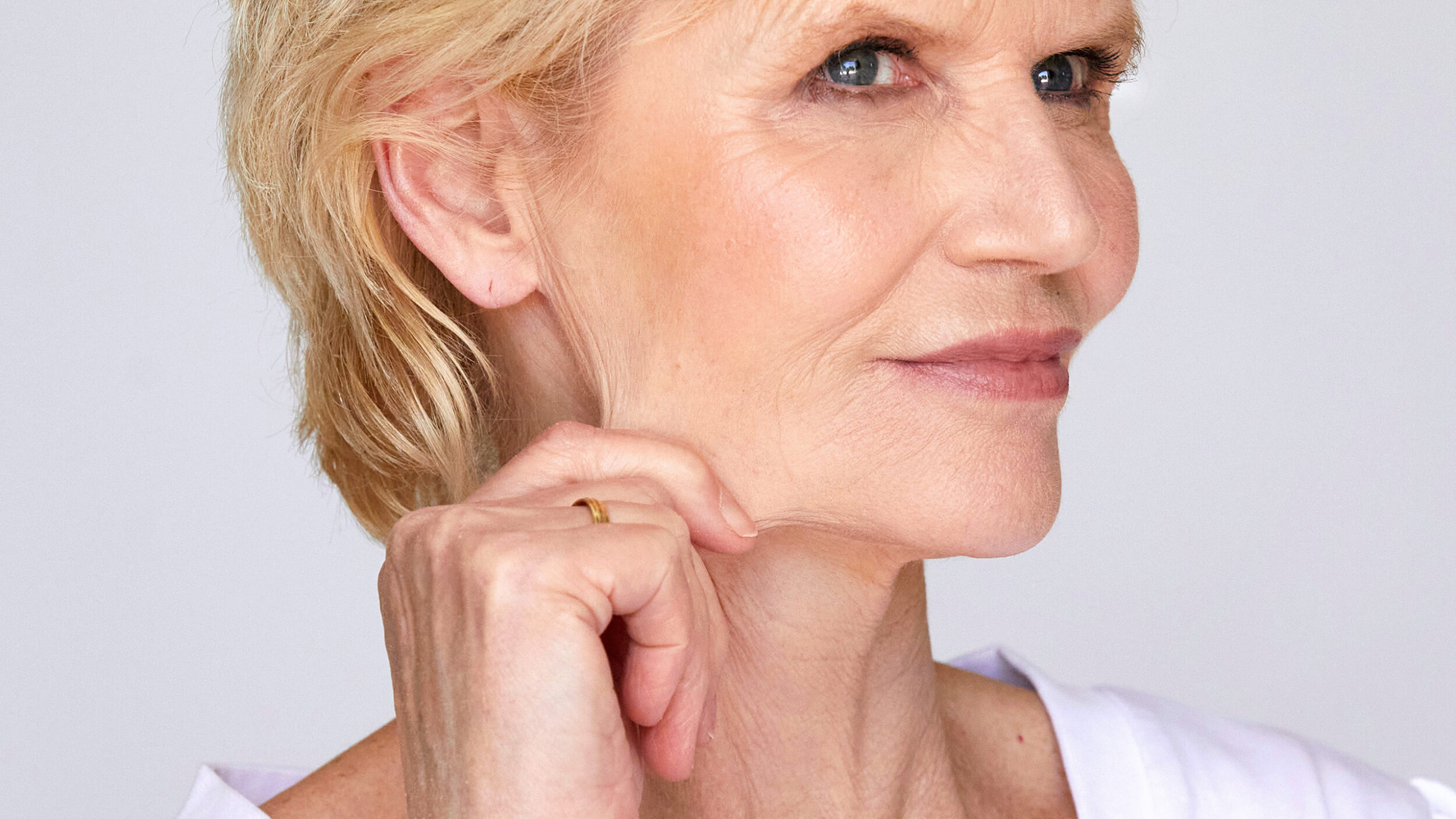 Mature woman gently lifting loose skin beneath the jaw after facial volume loss following weight loss.